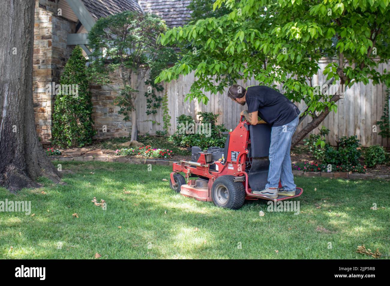 Tulsa USA 6 22 2018 Man with tattoos on standing commercial lawnmower cutting a close circle around tree and flowerbeds in upscale neighborhood - sele Stock Photo