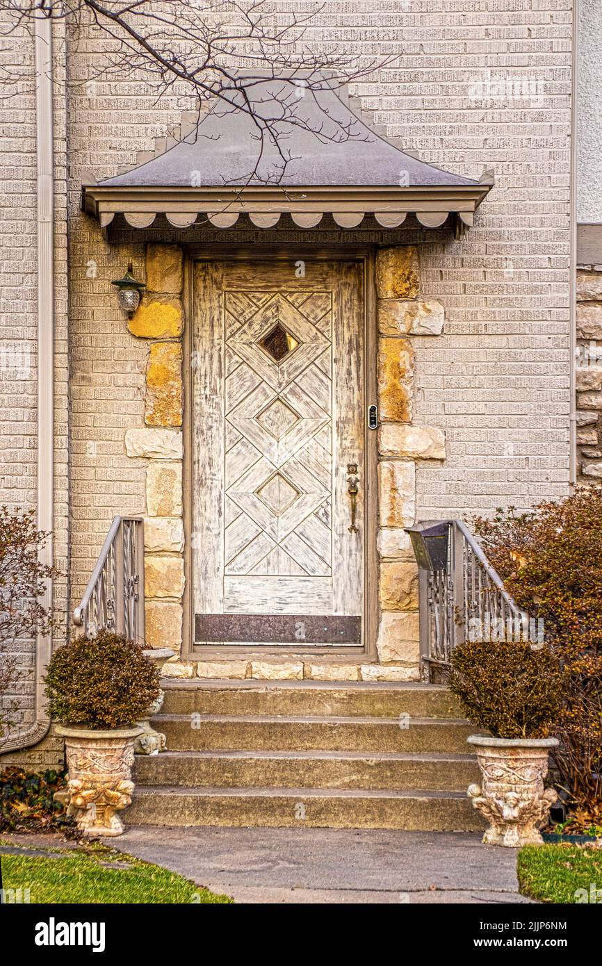 Unusual rustic wooden door surrounded by rock trim in light colored upscale brick house with awning on winter day with all bushes dead and brown Stock Photo