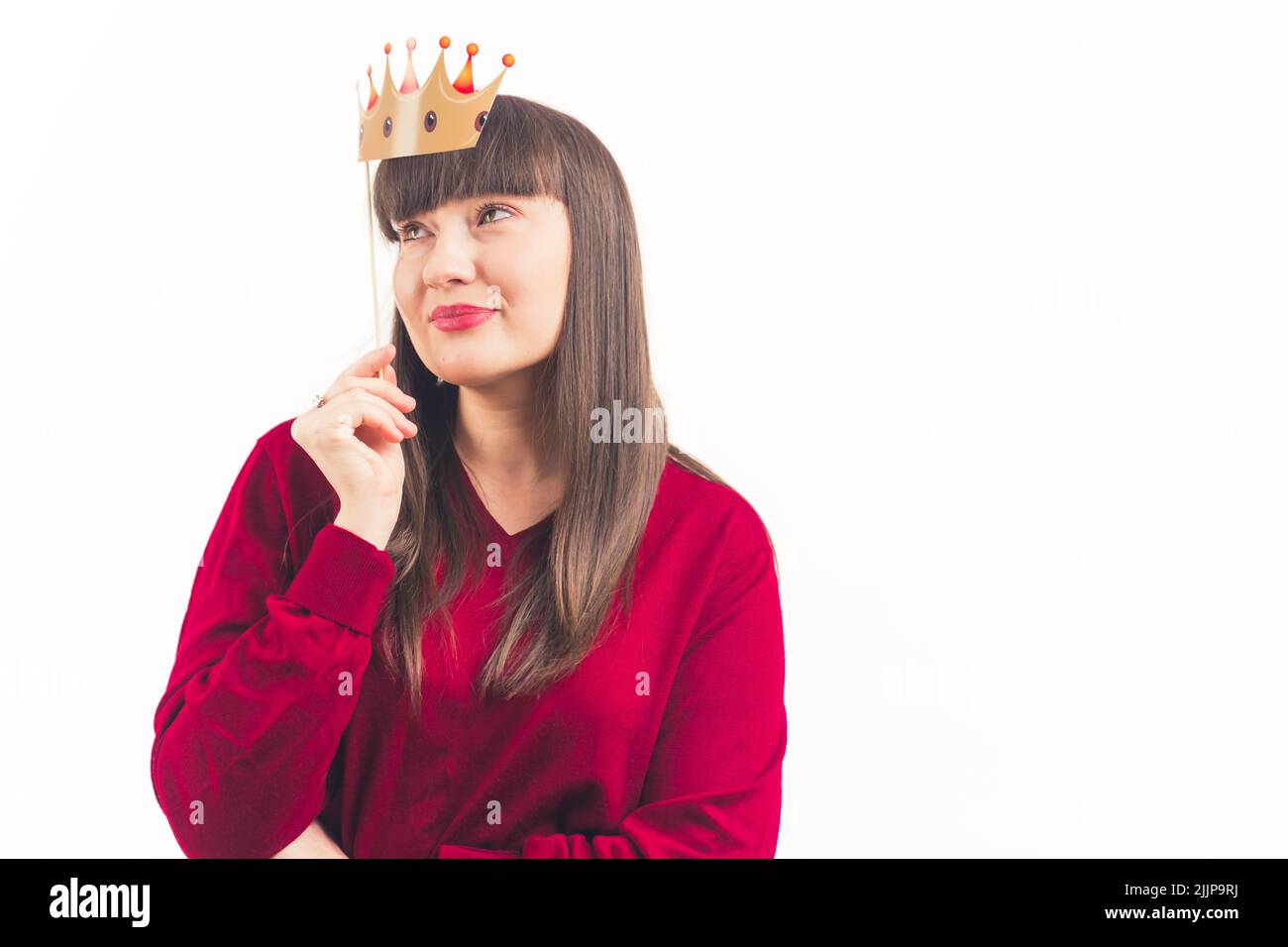 White dark-haired woman with bangs and soft makeup wearing red sweater holding fake paper crown above her face. Studio shot. Isolated copy space. High quality photo Stock Photo