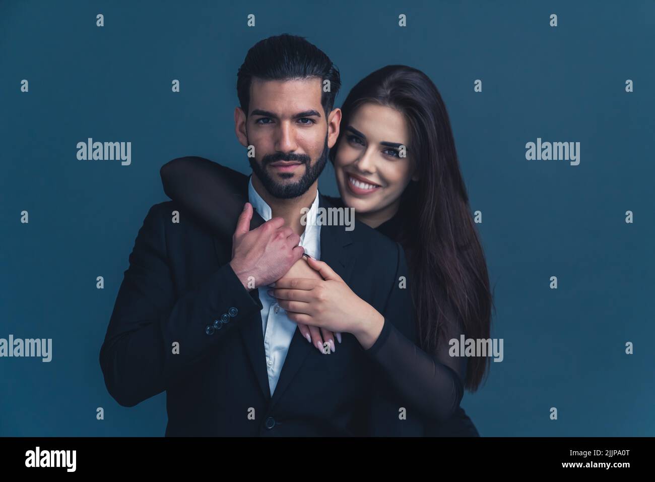 Handsome Cuban bearded guy in a black suit standing, looking at camera, and being held from the back by a beautiful long--haired caucasian girl with broad smile. Studio isolated shot. High quality photo Stock Photo