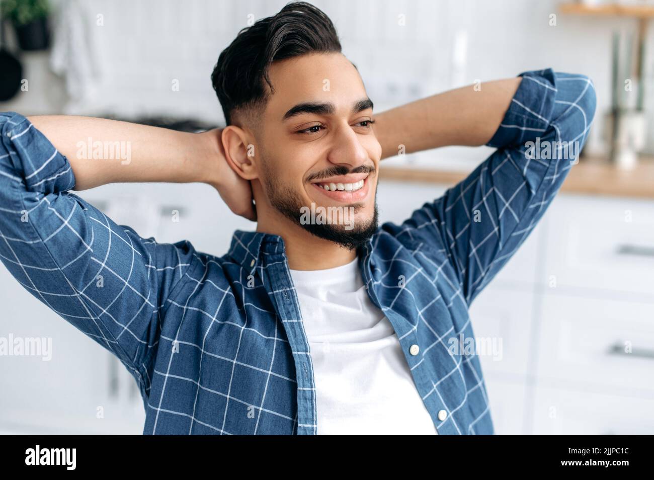 Close-up photo of a happy relaxed arabian or indian guy, freelancer, working from home, sits in the kitchen, takes a break from work, puts his hands behind his head, looks directly, dreaming, smiles Stock Photo