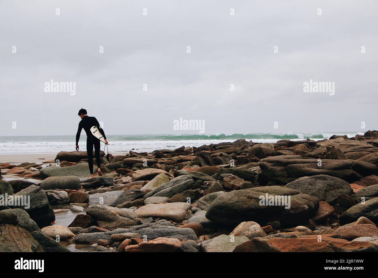 A young surfer walking across a rocky shore to the sea.  A picture processed with VSCO with a6 preset Stock Photo