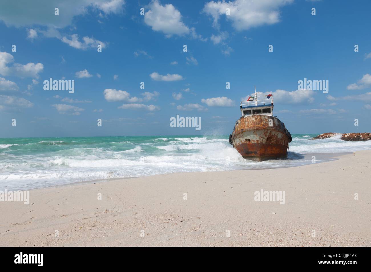 Stranded rusty fisherman boat photographed at the coast of Ajman Emirate on sunny winter day. Sea, sand, blue sky with clouds at background. Stock Photo