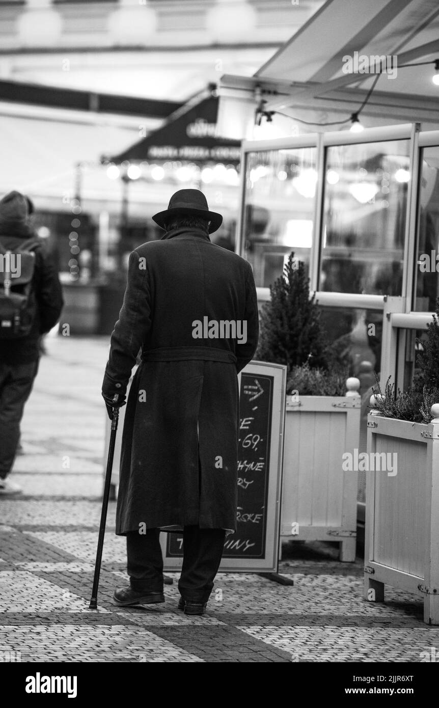 A grayscale vertical shot of an old man wearing a coat and hat and walking with a stick on a street Stock Photo