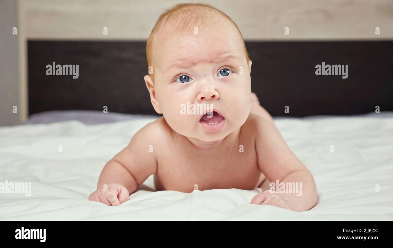 Cute baby girl frowns trying to raise up head lying on bed Stock Photo
