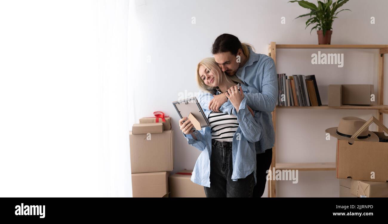 young couple move at new home standing take out unpack belongings from carton boxes holds frame looks at photo share memories feels happy. Buy first Stock Photo
