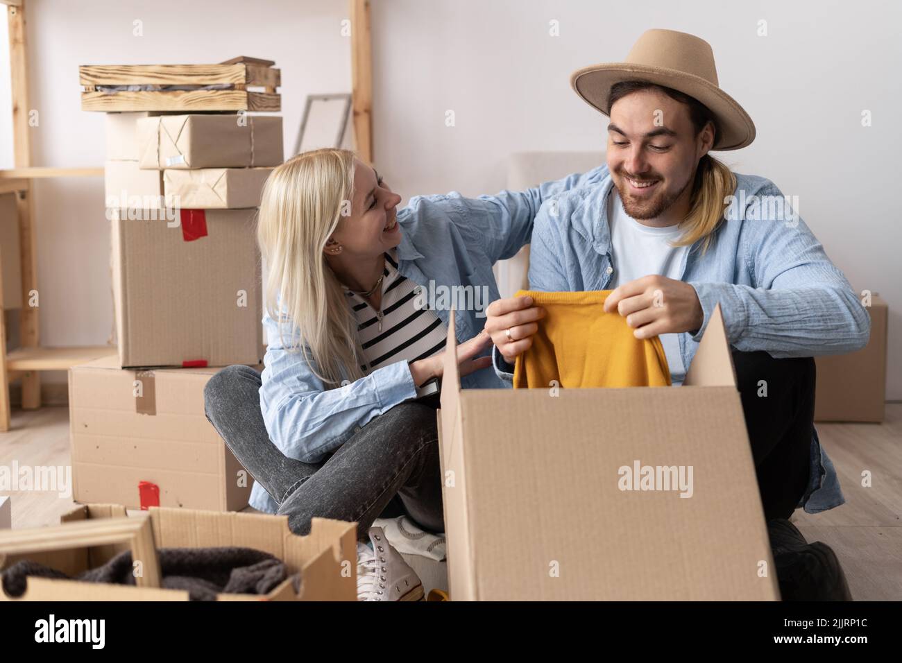 a young cheerful couple in a new apartment on the day of the move sit on the floor unpack things the woman puts on a hat to the man. moving to a new Stock Photo