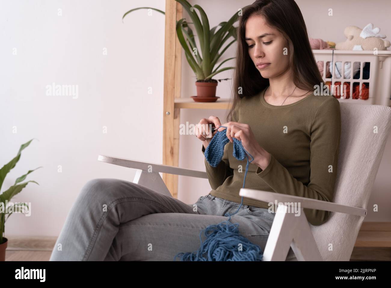 latin young woman knitting while sitting on armchair at home, hobby and needlework, handmade knitwear, leisure and hobby Stock Photo