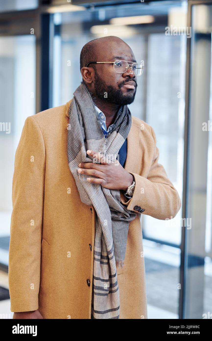 African confident businessman in elegant coat putting on the scarf around his neck, he leaving the office building Stock Photo
