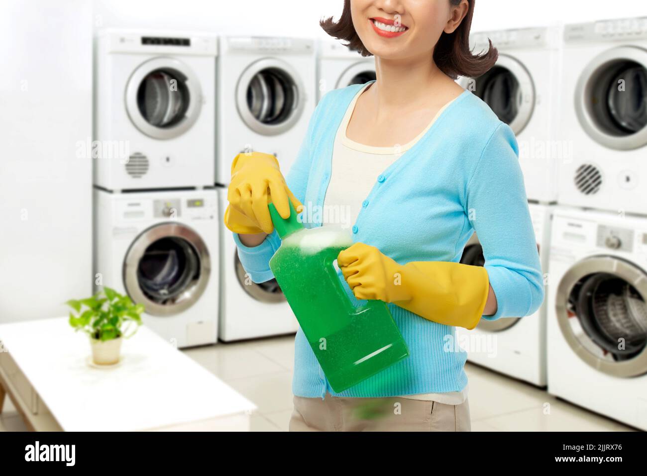 Smiling young woman wearing rubber gloves opening bottle of liquid detergent while standing at spacious self-service laundry Stock Photo