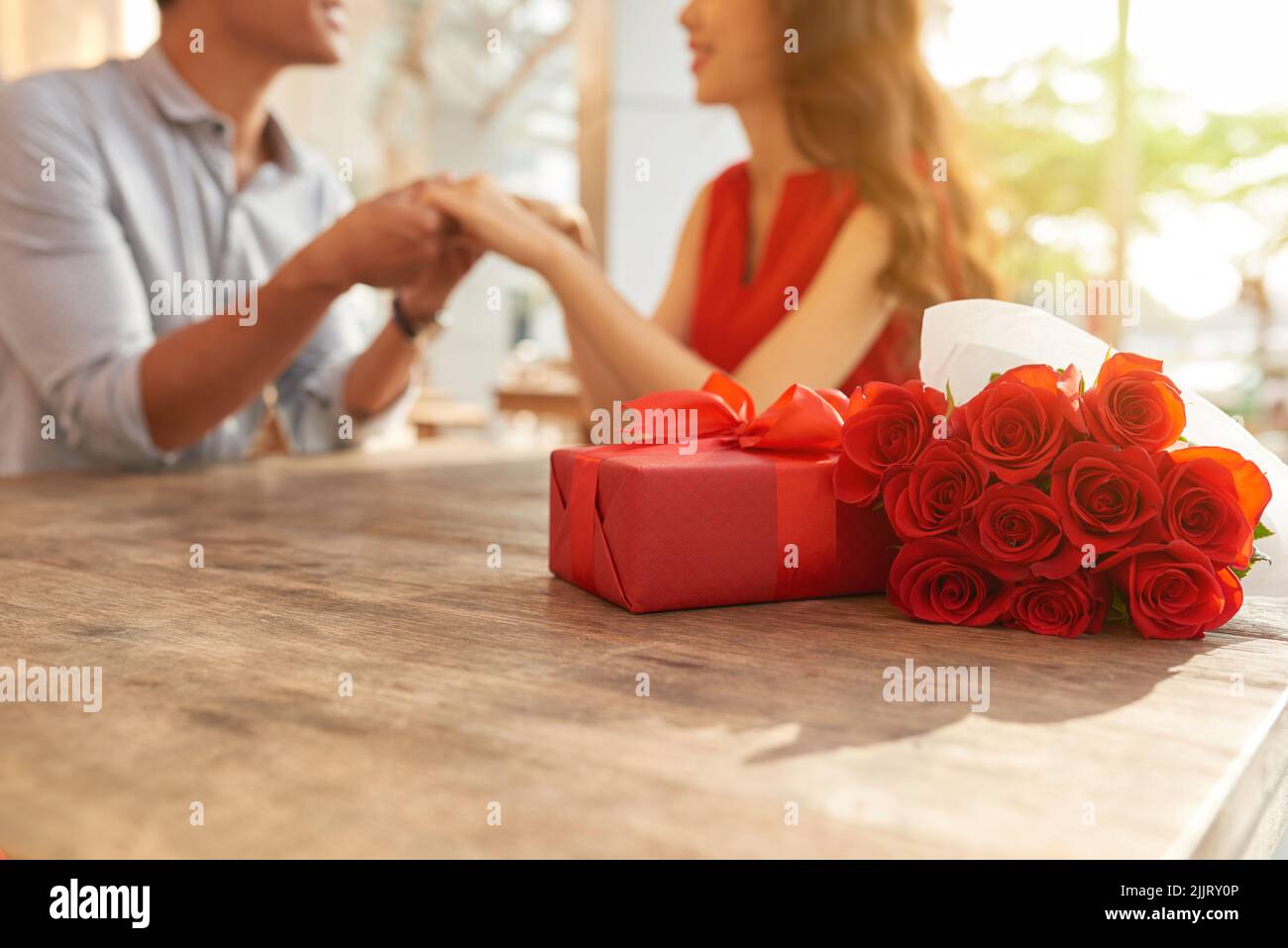 Cheerful young couple sitting at wooden table of outdoor cafe and holding hands while celebrating Valentines Day, gift box and bouquet of red roses on foreground Stock Photo