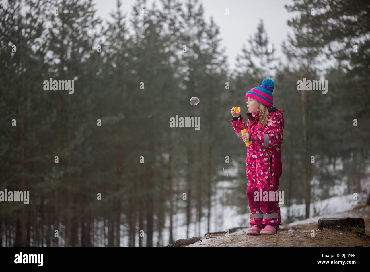 A cute Caucasian baby girl in a pink winter overall blowing bubbles in the snowy forest Stock Photo