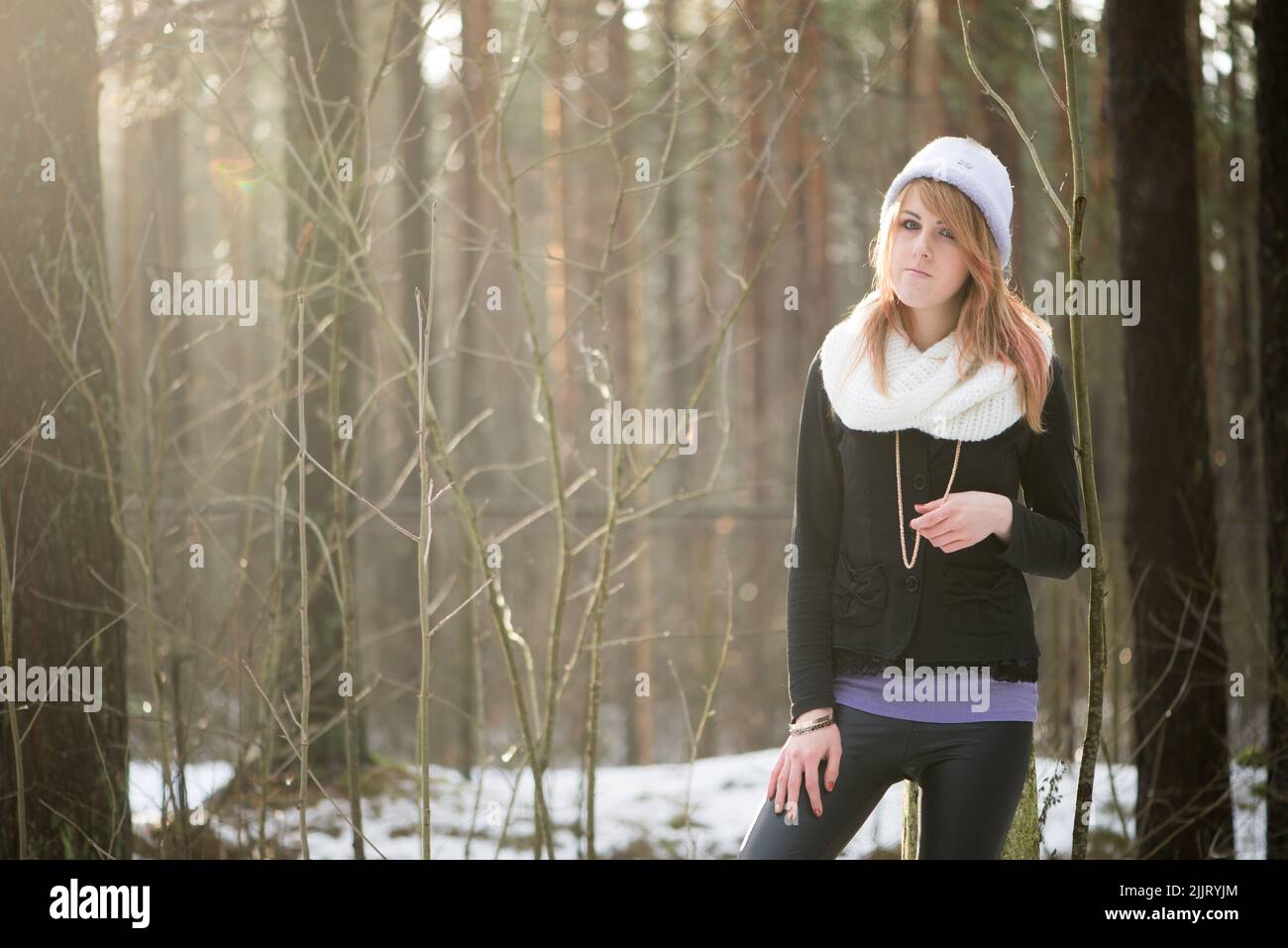 A Caucasian blonde girl in a winter jacket and hat posing with a blurred background of leafless trees Stock Photo