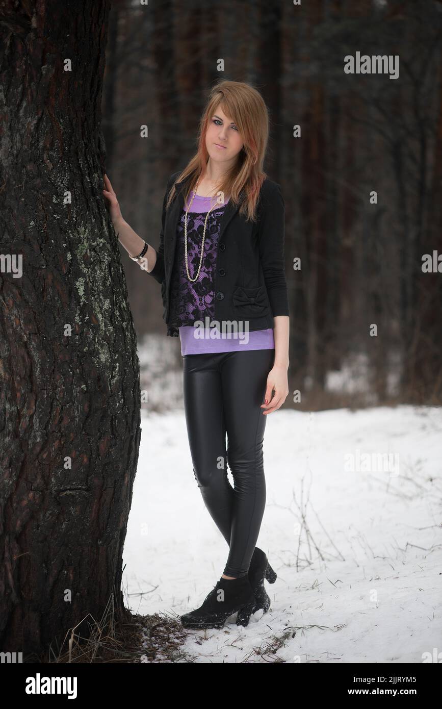 A Caucasian blonde girl in a winter jacket and hat posing with a blurred background of leafless trees Stock Photo