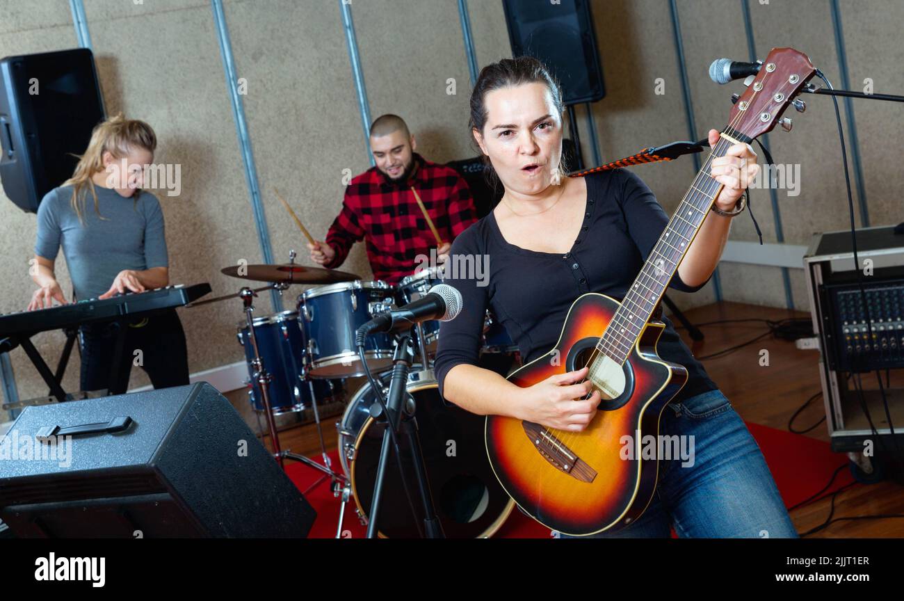 Attractive female soloist playing guitar and singing with her music band in sound studio Stock Photo