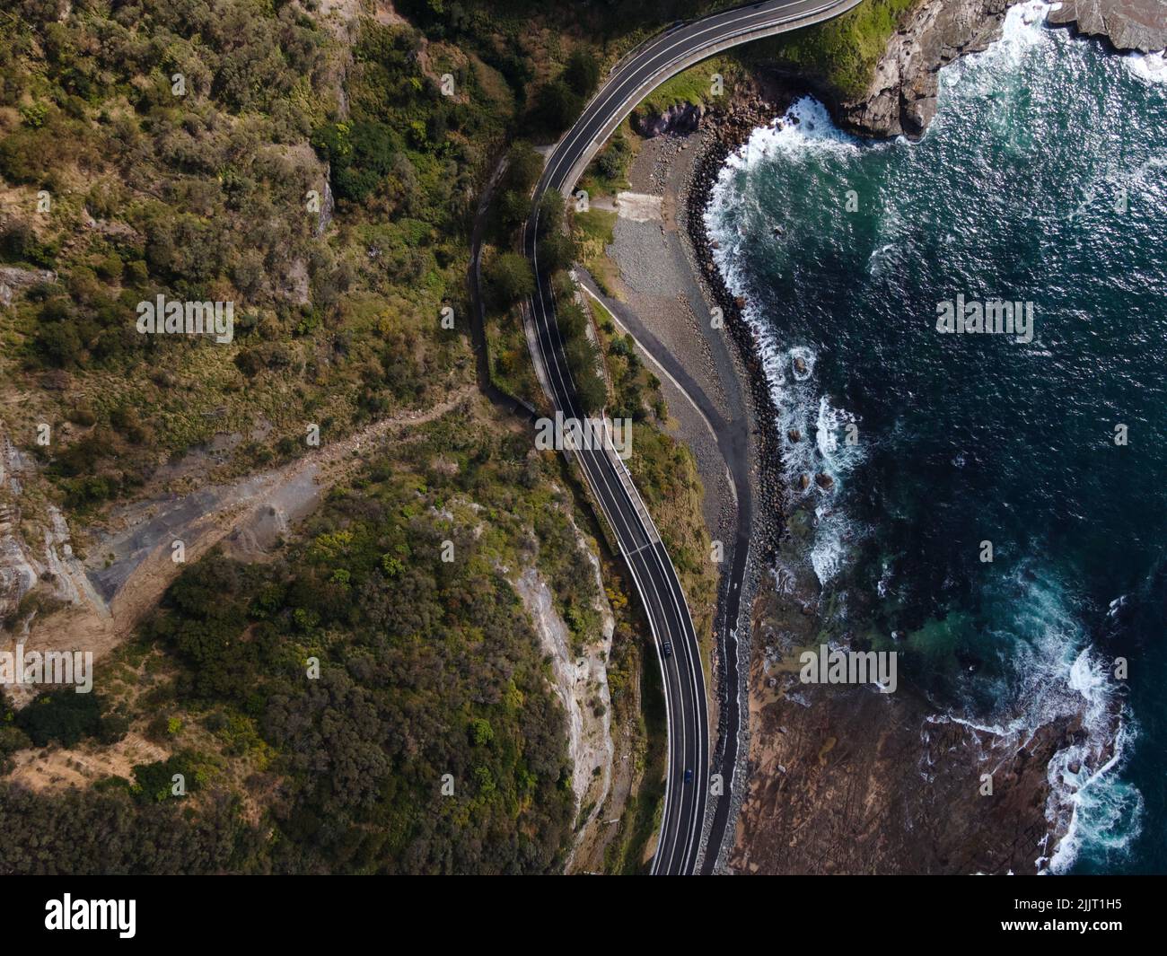 Windy ocean road next to the water and around rocks and trees Stock Photo