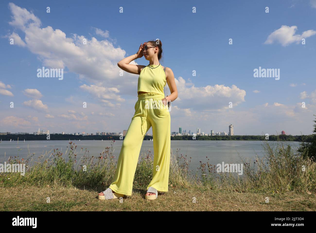 Slim girl in sunglasses and yellow summer suit standing on hill top on background of water and city buildings. Females fashion, travel and holidays Stock Photo