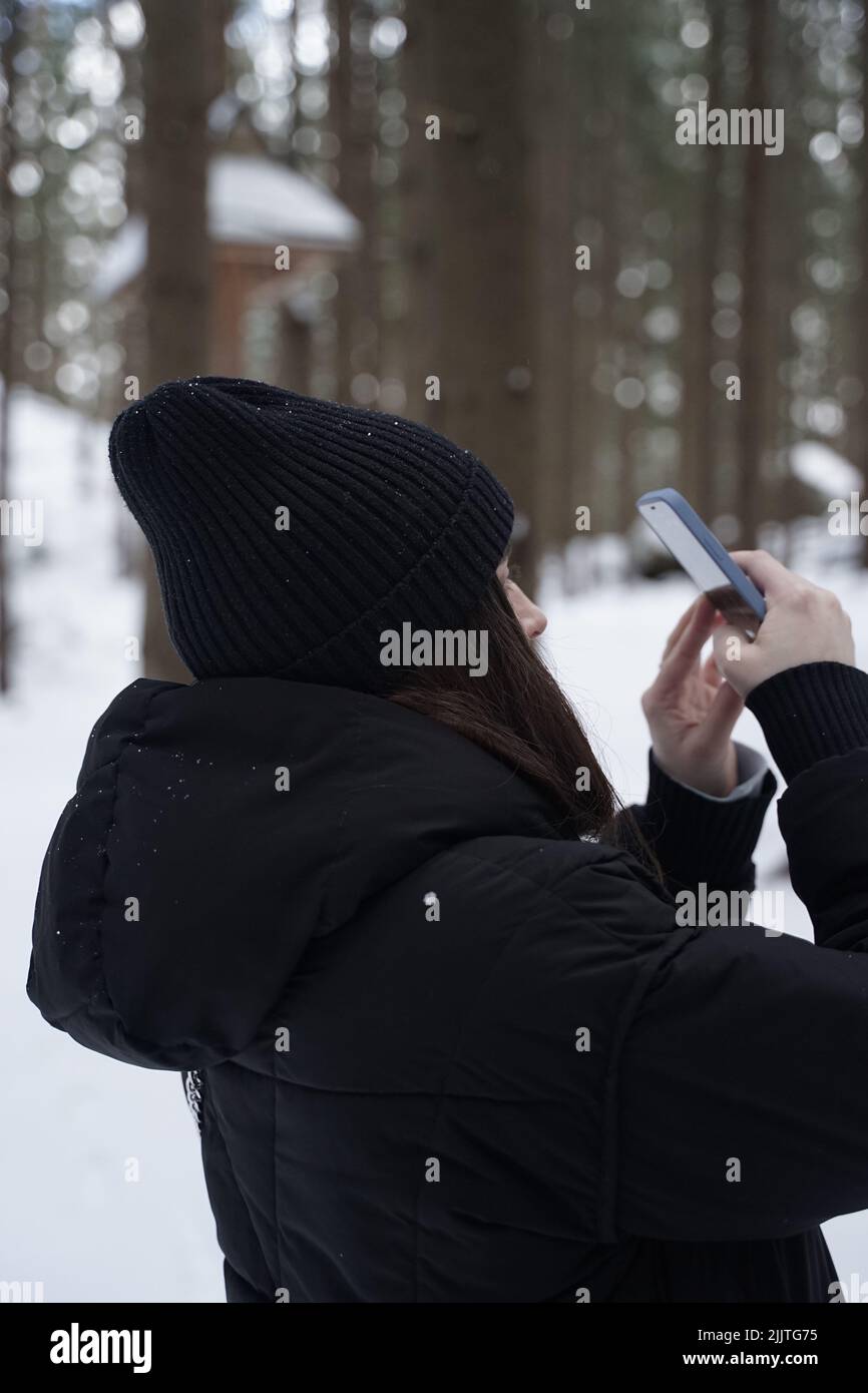 A vertical shot of a caucasian female taking pictures on her smartphone in a winter forest Stock Photo