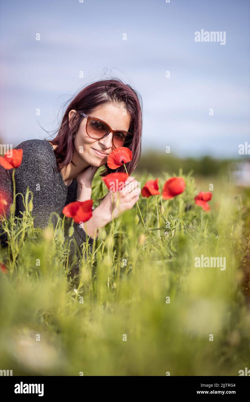 A beautiful portrait of a young Caucasian woman sniffing a red flower in the field in bright sunlight Stock Photo