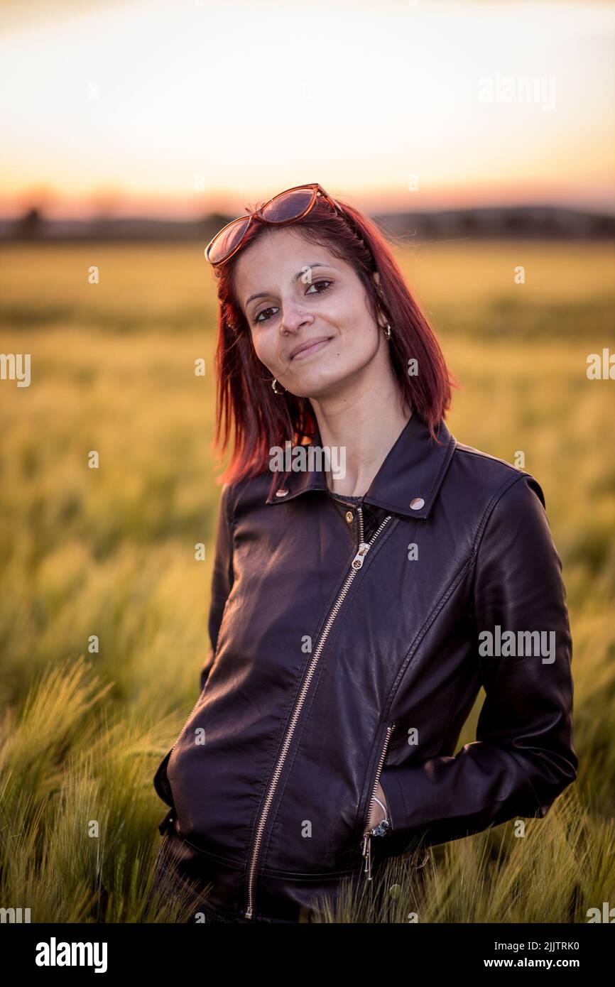 A vertical closeup of a young woman in a leather jacket standing in the green field looking at camera. Stock Photo