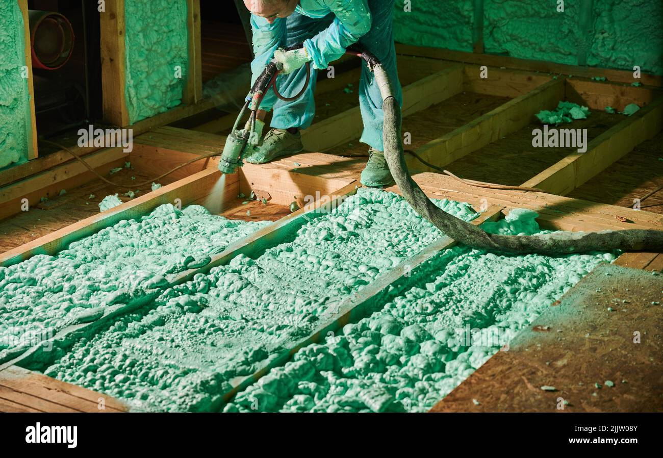 Male builder insulating wooden frame house. Cropped view of man worker spraying polyurethane foam on floor inside of future cottage, using plural component gun. Construction and insulation concept. Stock Photo