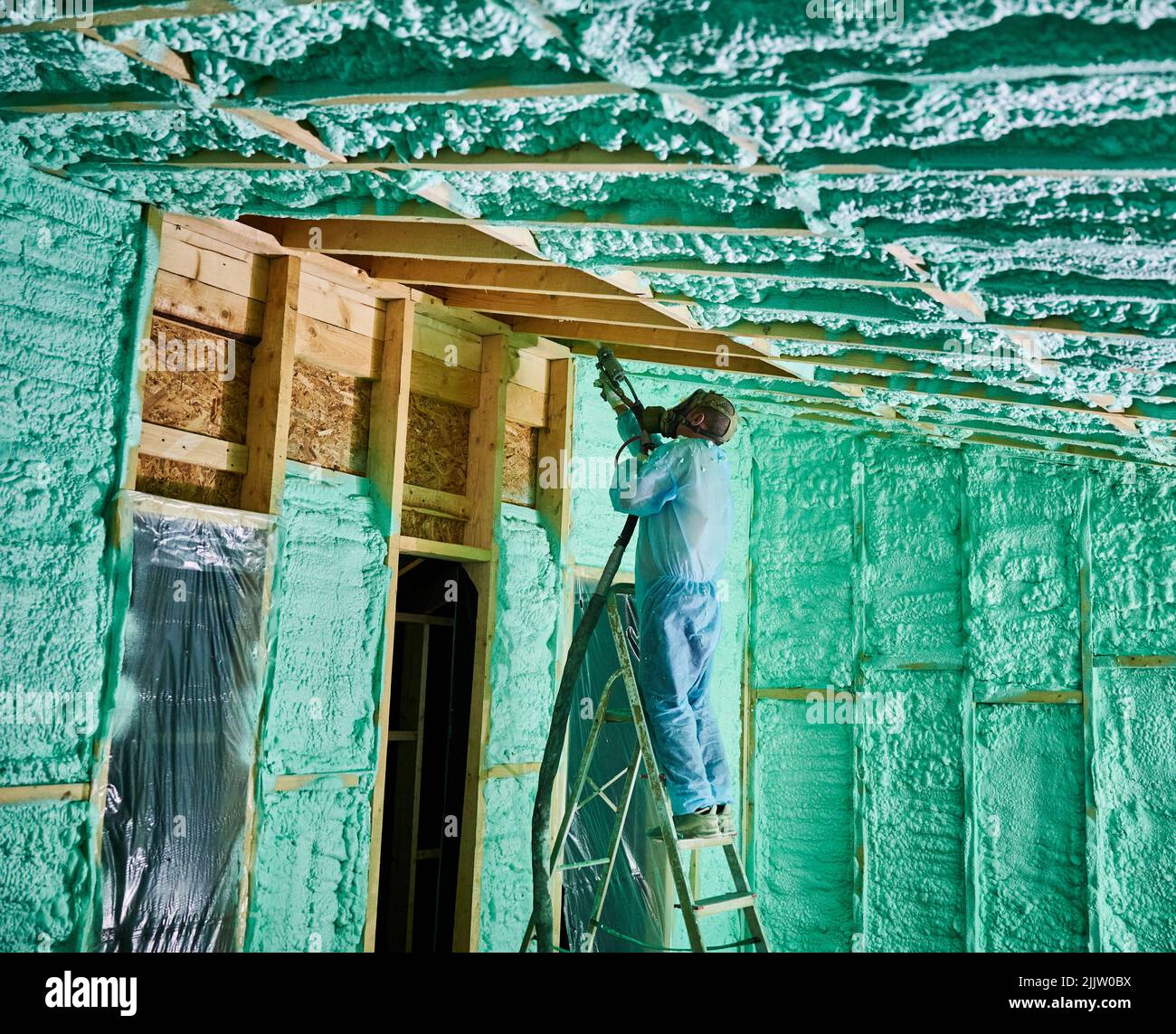 Male builder insulating wooden frame house. Man worker spraying polyurethane foam inside of future cottage, standing on ladder, using plural component gun. Construction and insulation concept. Stock Photo