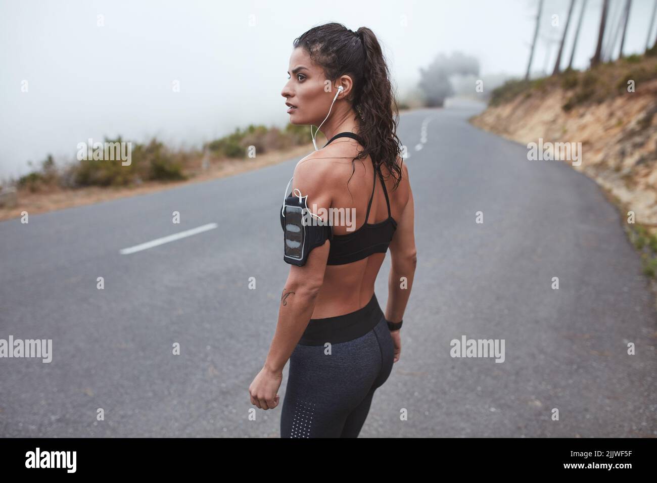 Fit young woman going out for a morning run along a misty road. Sporty young woman listening to music during her training session. Stock Photo