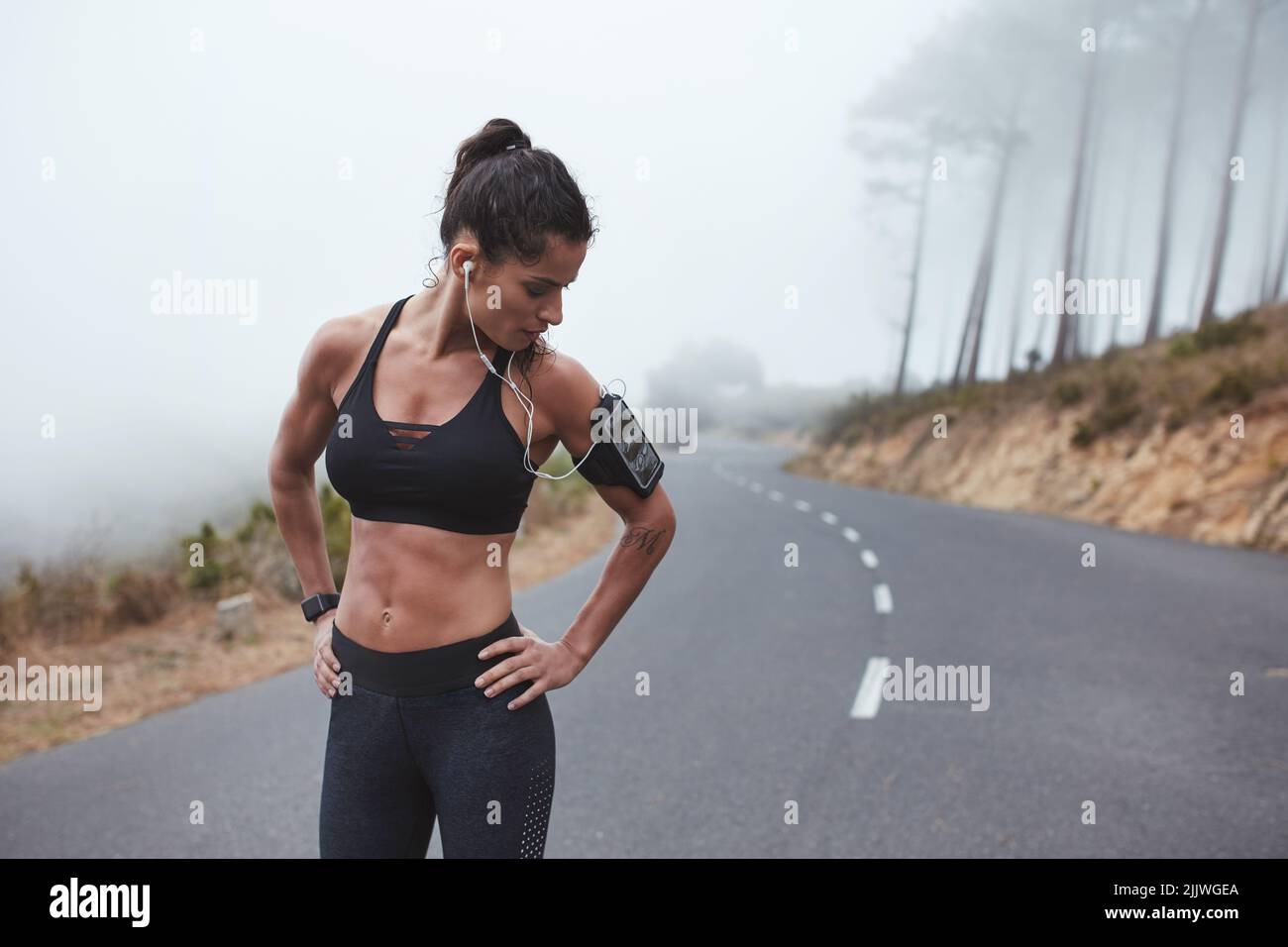 Female athlete looking at her smartphone while standing on a misty road in sportswear. Fit young woman listening to music during her morning training Stock Photo