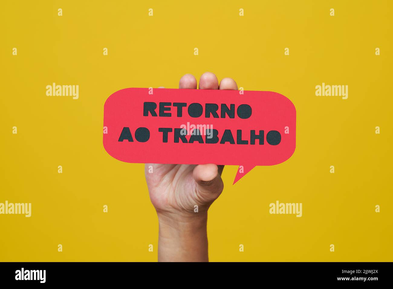 a man holds a red paper sign, in the shape of a speech bubble, that reads the text back to work written in portuguese, on a yellow background Stock Photo