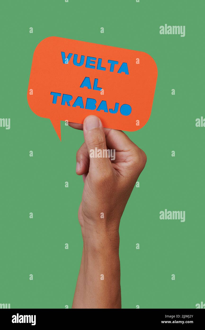 man holds an orange paper sign, in the shape of a speech bubble, that reads the text back to work written in spanish, on a green background Stock Photo