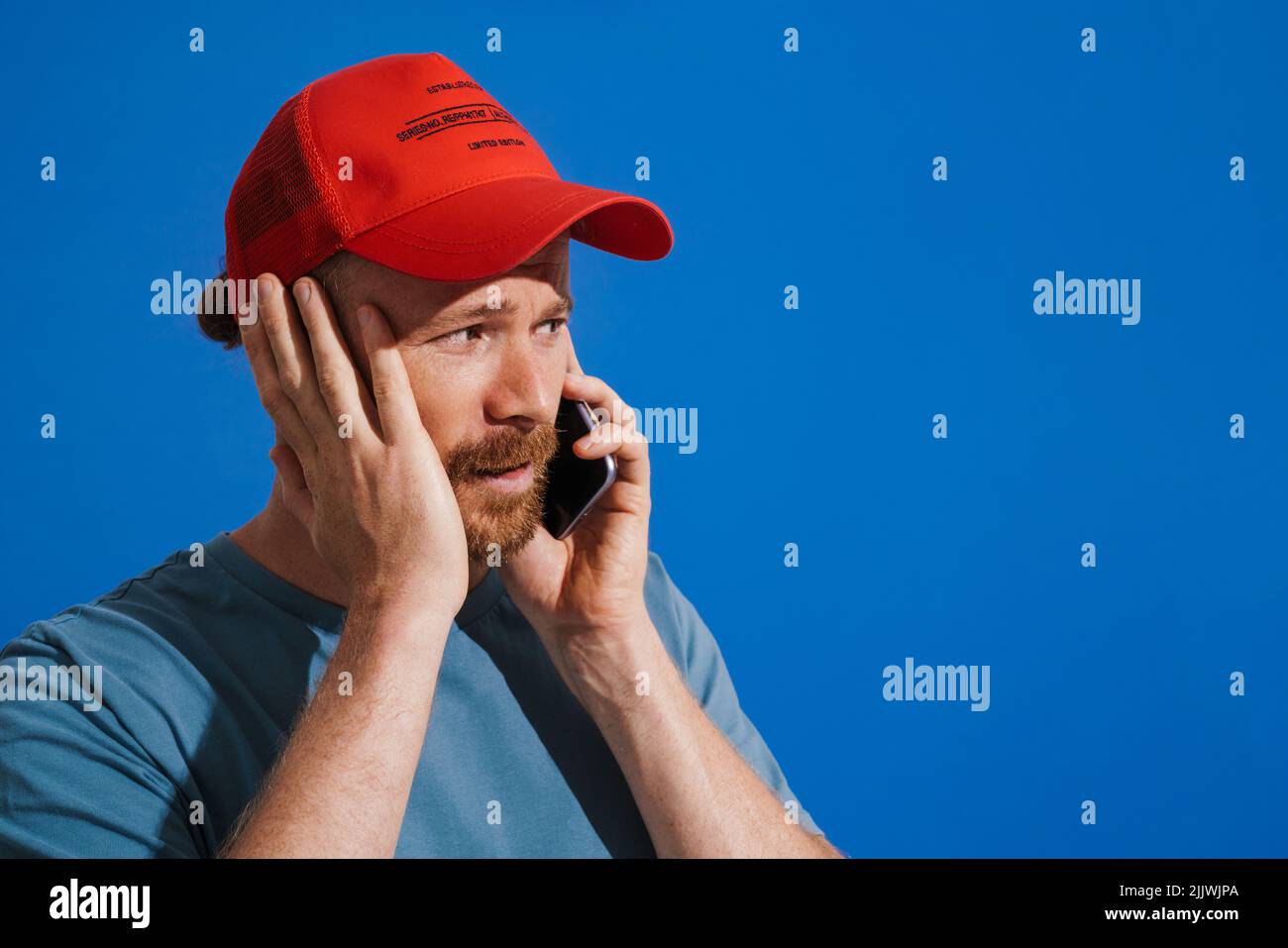 White ginger man wearing cap talking on mobile phone isolated over blue background Stock Photo