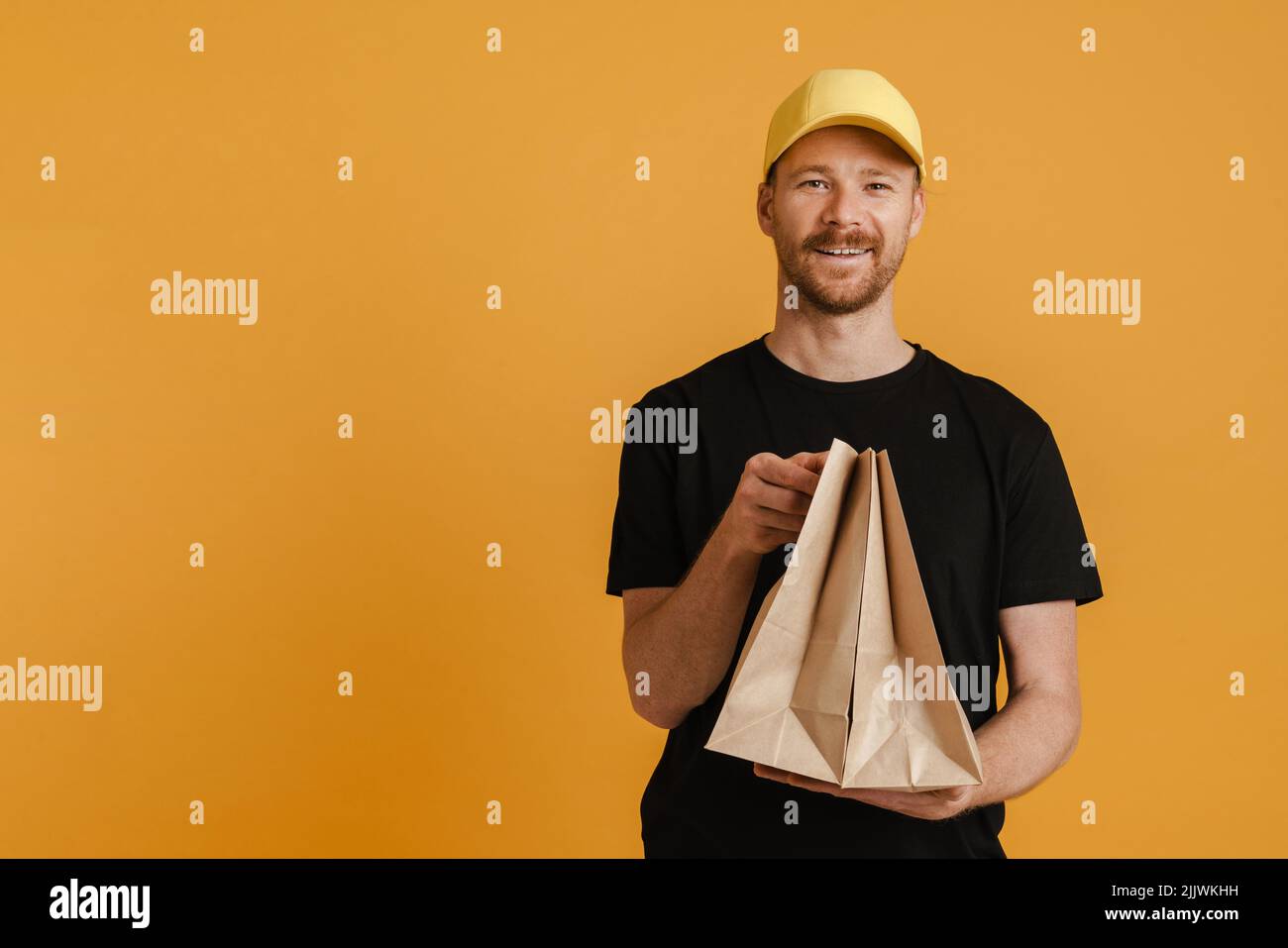 White delivery man in cap smiling while posing with paper bags isolated over yellow background Stock Photo