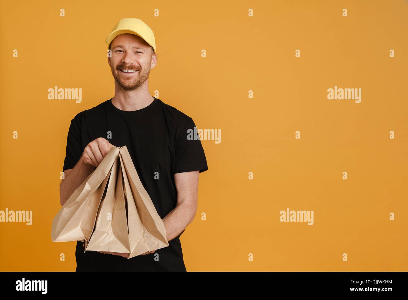 White delivery man in cap smiling while posing with paper bags isolated over yellow background Stock Photo