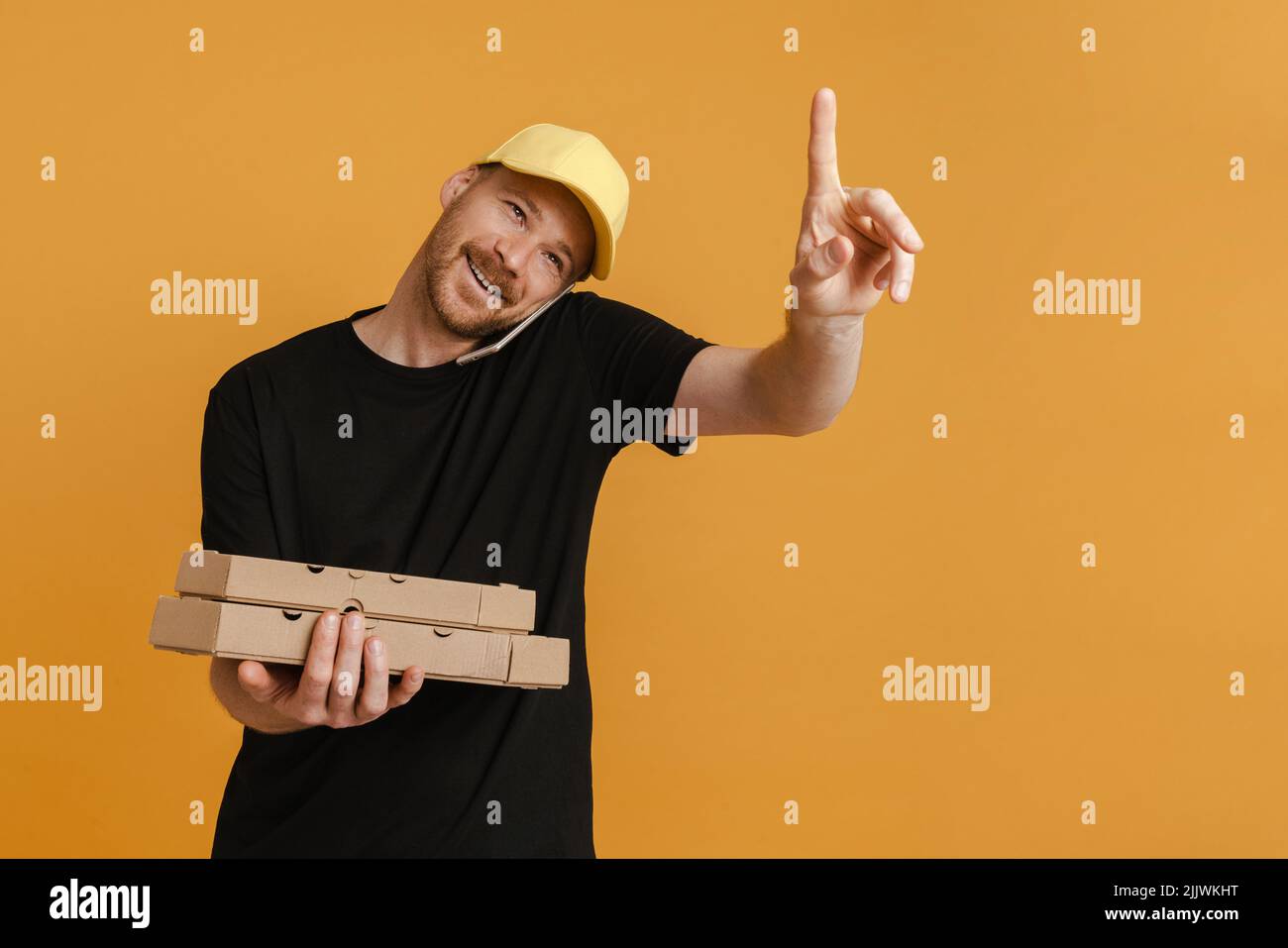 White delivery man with pizza boxes talking on mobile phone isolated over yellow background Stock Photo