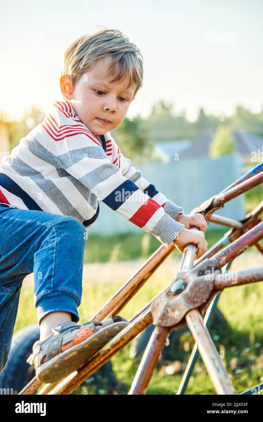 Cute preschooler holds on to old rusty climbing play set on sunny day on blurred background. Little blond boy spends summer vacation in countryside Stock Photo