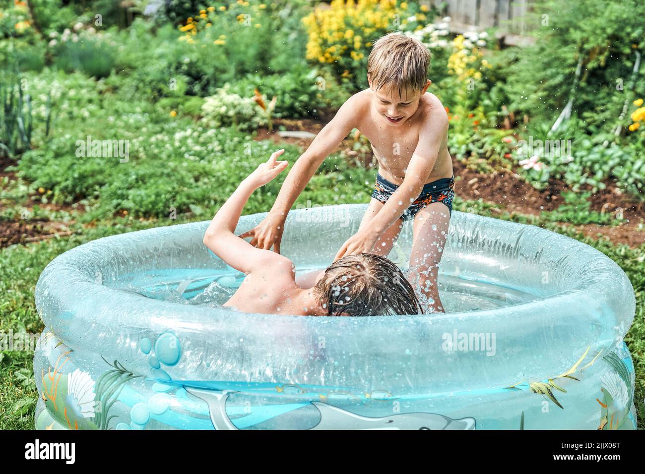Brothers wrestle in children inflatable pool in cottage yard against lush greenery and flowers. Cheerful schoolboys have fun in water on summer in cou Stock Photo