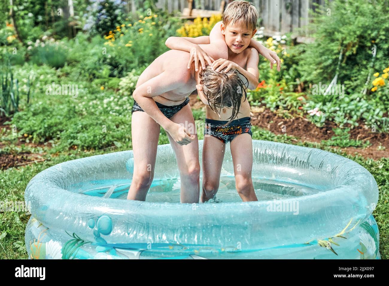 Brothers wrestle in children inflatable pool in cottage yard against lush greenery and flowers. Cheerful schoolboys have fun in water on summer in cou Stock Photo