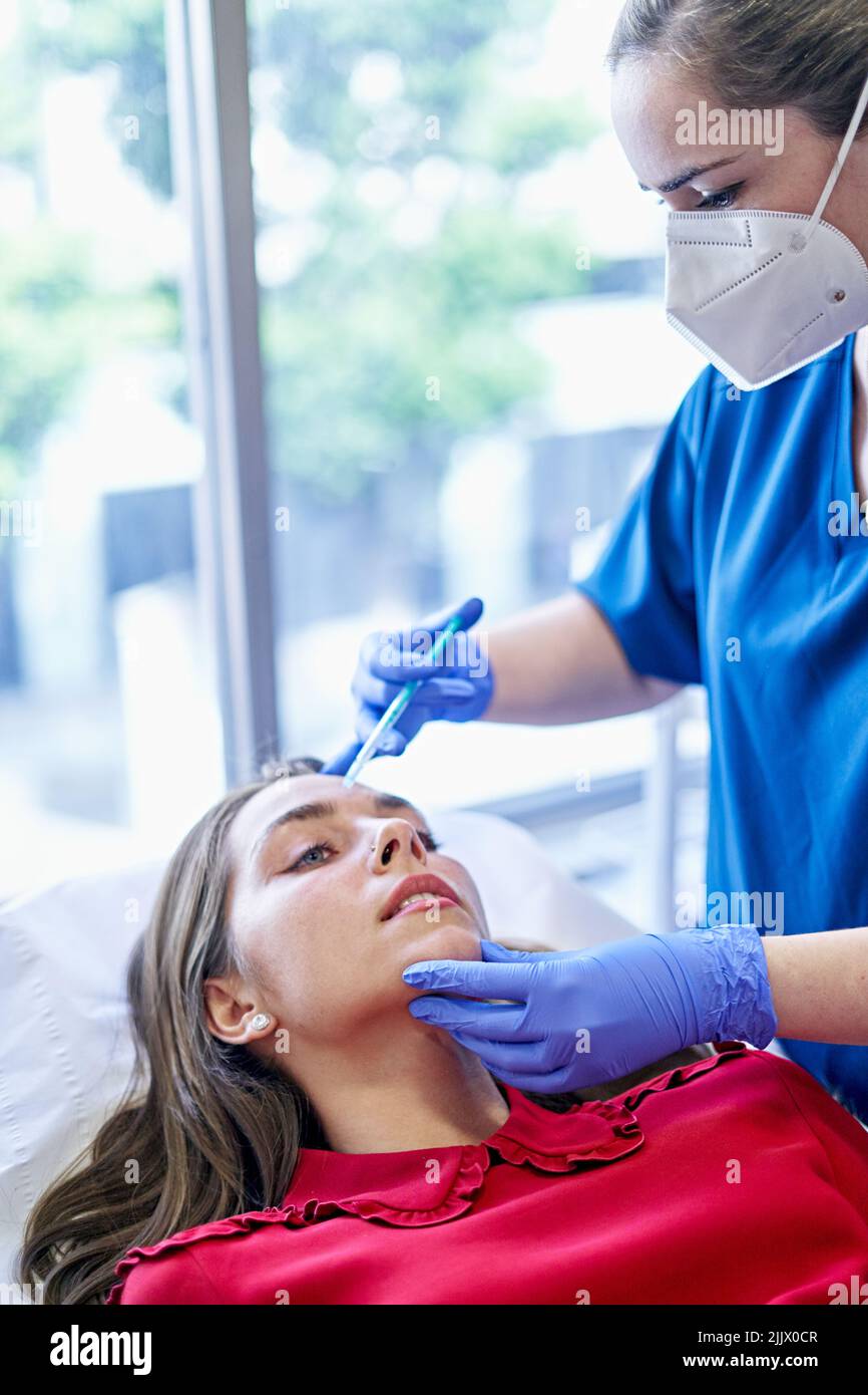 Esthetician in uniform and face mask injecting collagen into forehead ...