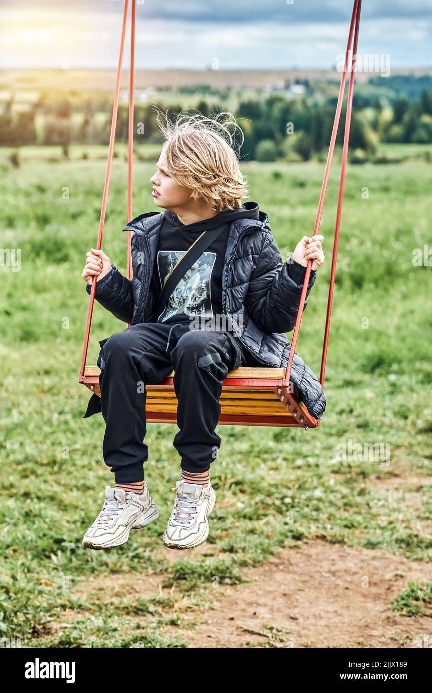 Cute schoolboy with long blond hair rides on swing looking aside. Boy enjoys nature against green grass and lush trees on cloudy day in countryside. N Stock Photo