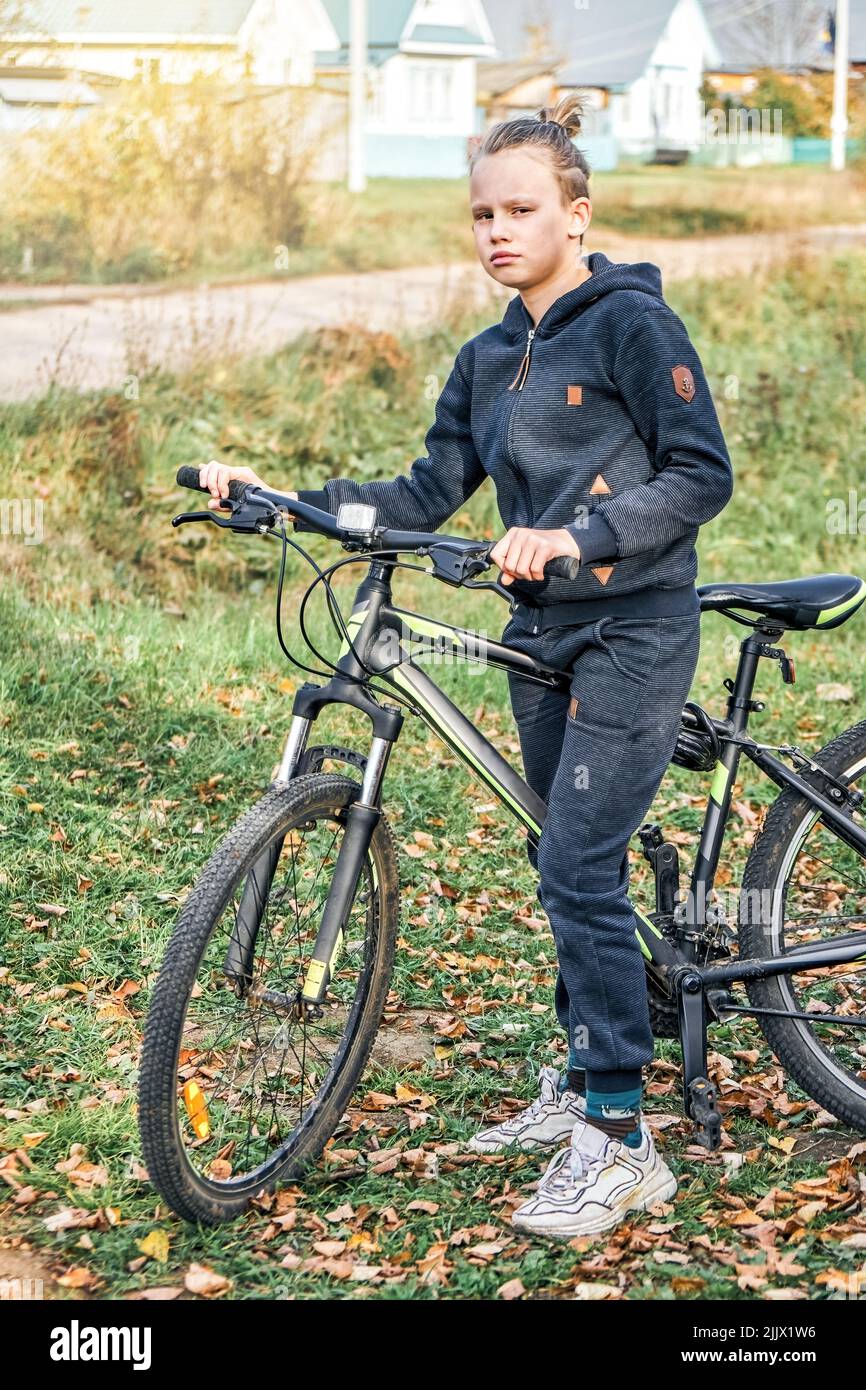 Blond junior schoolboy in casual outfit enjoys riding bicycle in countryside. Boy stands on green grass against houses near road on autumn day Stock Photo