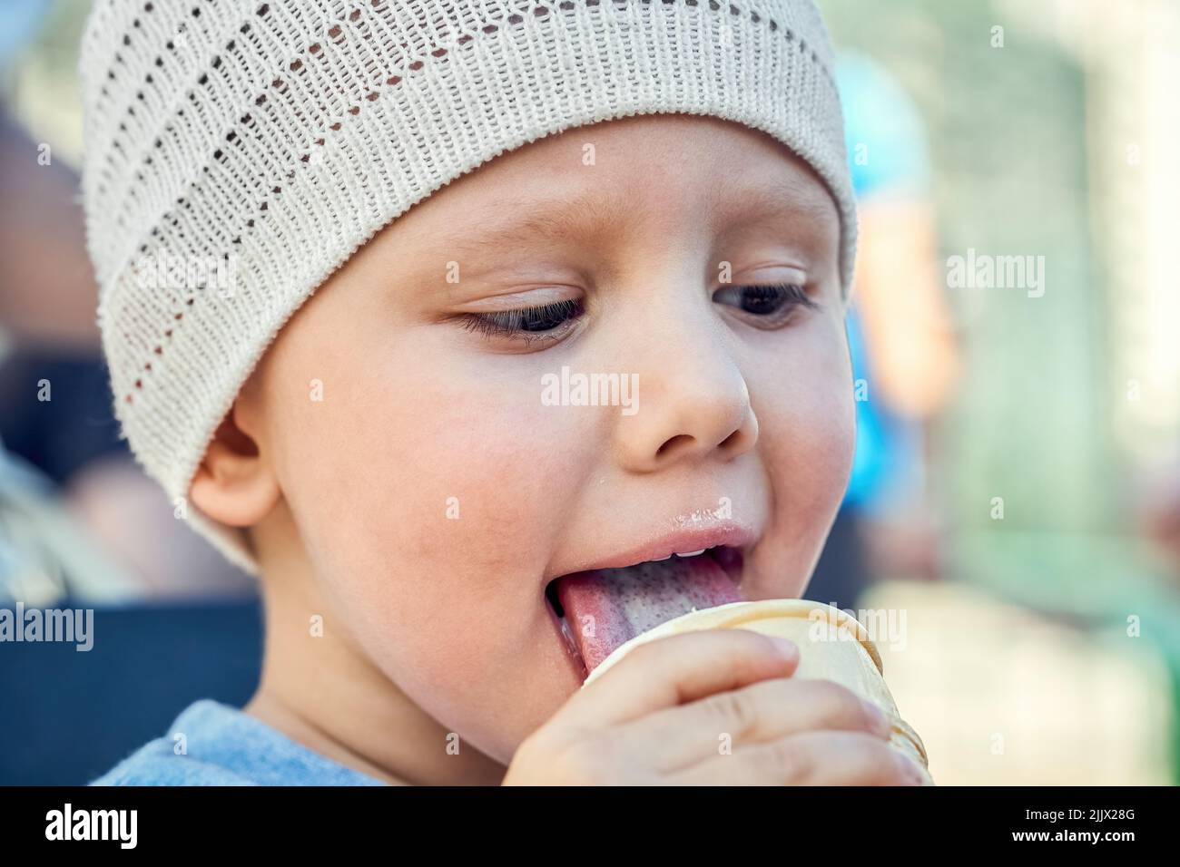 Little boy in white knitted hat licks ice cream and looks down. Toddler enjoys milk ice-cream in waffle cup closeup. Happy carefree childhood concept Stock Photo