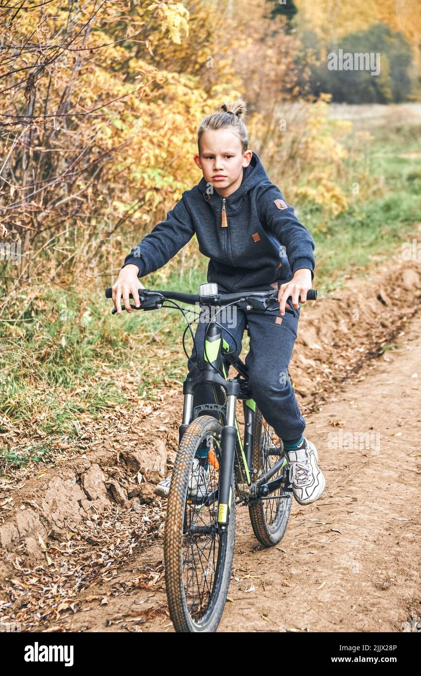 Blond boy in casual outfit rides bicycle on ground road past trees with yellowed leaves in countryside. Schoolboy enjoys sports activity on autumn day Stock Photo
