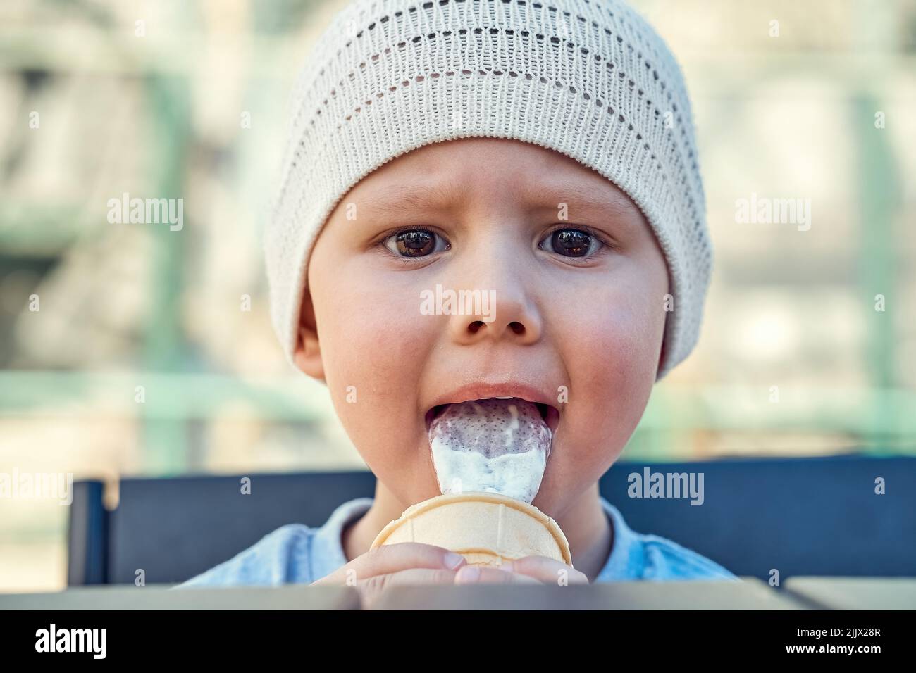 Toddler boy eats ice cream with pleasure and looks at camera closeup. Boy in white knitted hat licks ice-cream sitting on bench on playground. Walk in Stock Photo
