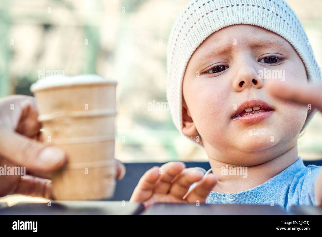 Dad feeds focused toddler with ice cream on playground bench. Boy in white knitted hat sits at table looking aside with interest closeup Stock Photo