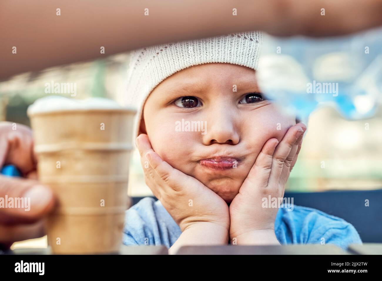 Small boy in white knitted hat sits at table with water in mouth holding cheeks with hands. Parent gives water to toddler holding ice cream in hand cl Stock Photo