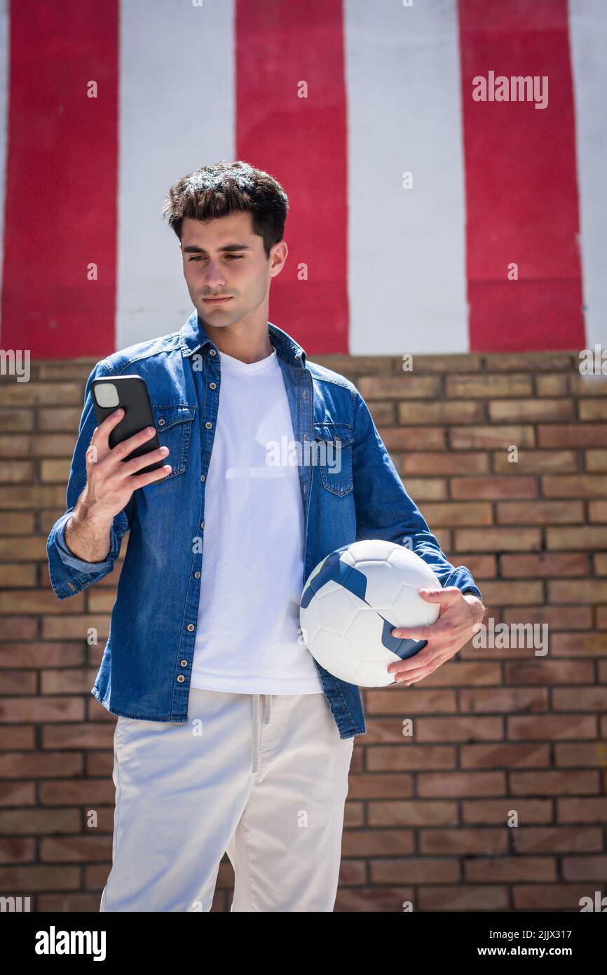 Young man with football using smartphone standing near brick wall Stock Photo