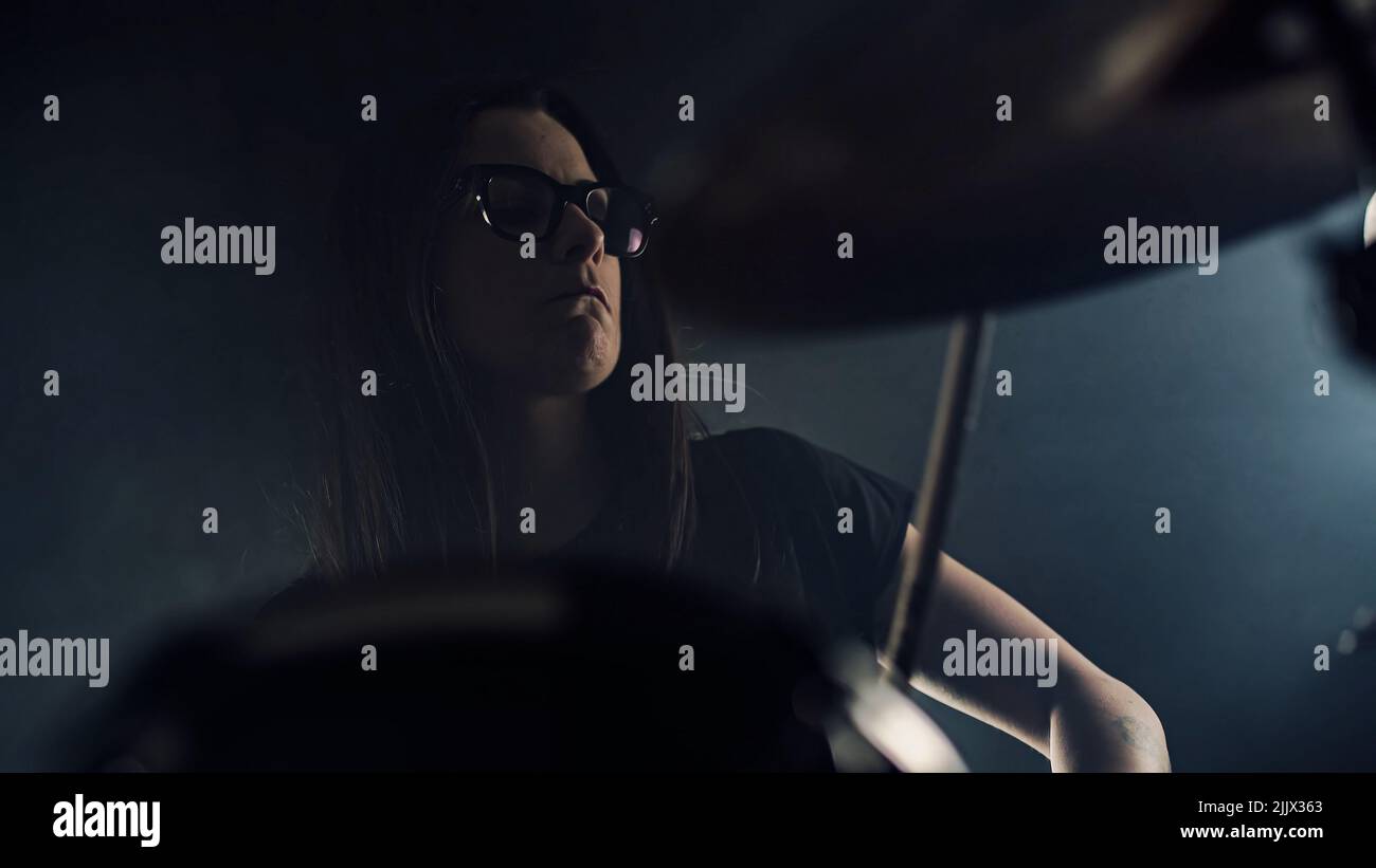 Young female drummer with spectacles plays a drum set holding drumstick in hands Stock Photo