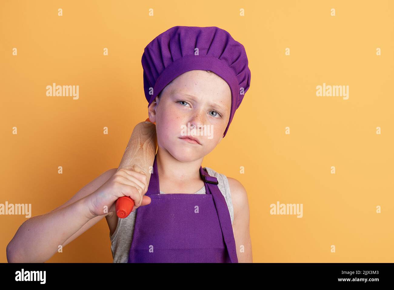 Serious little child in purple chef apron and hat putting rolling pin