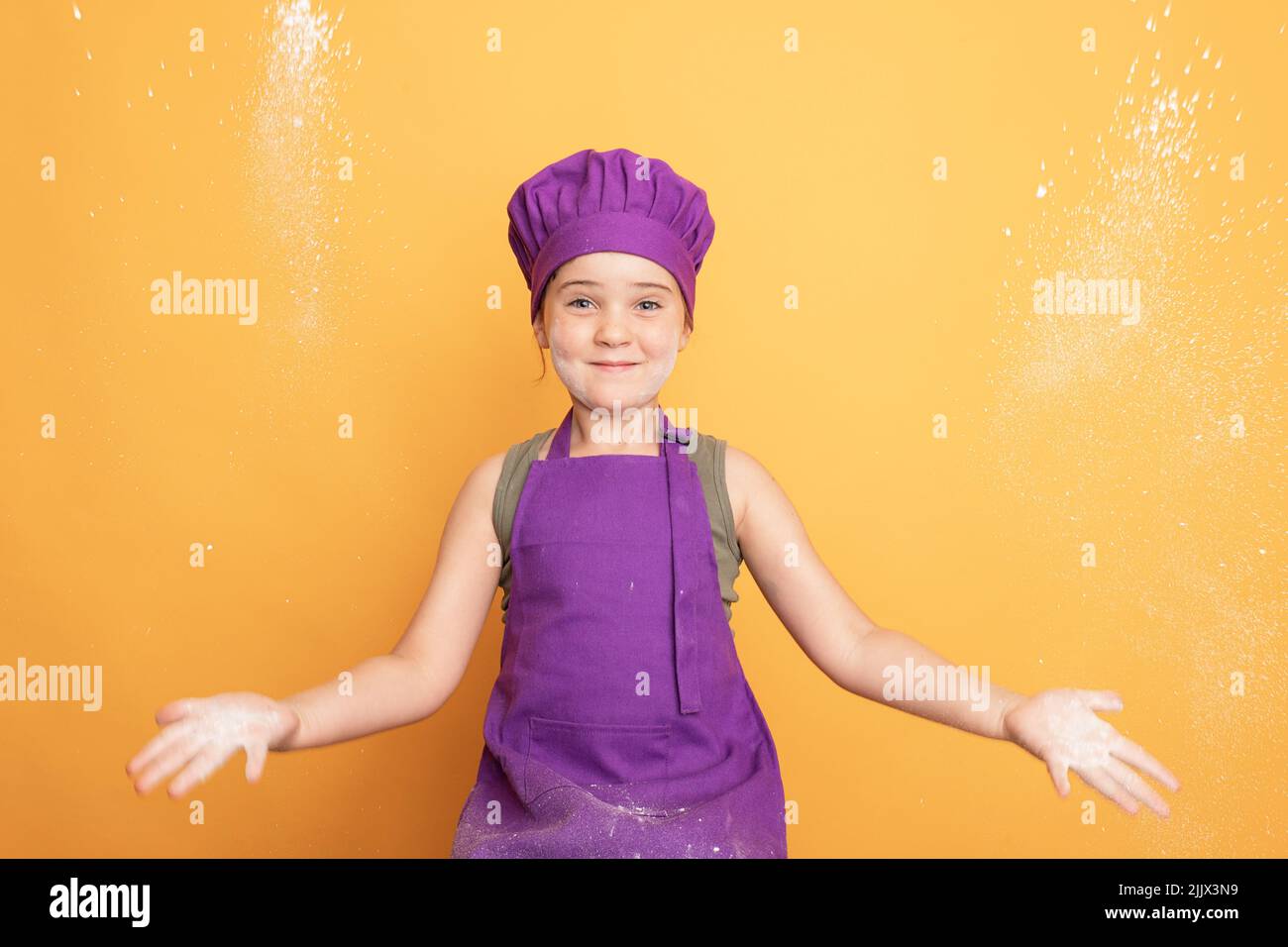 Optimistic girl in purple chef uniform clapping hands with white flour