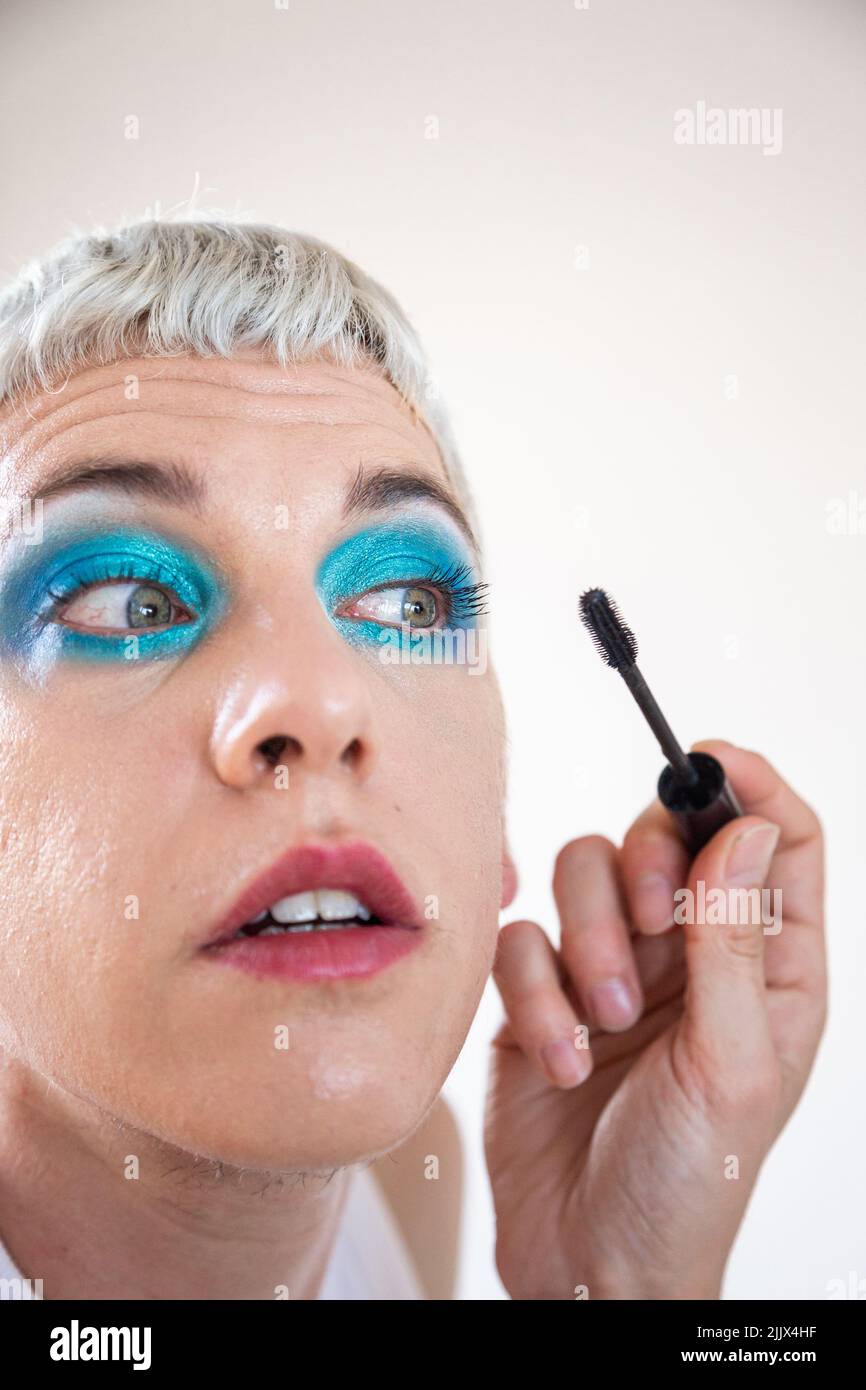 Closeup of transgender man wearing blue eyeshadow applying mascara on white background Stock Photo